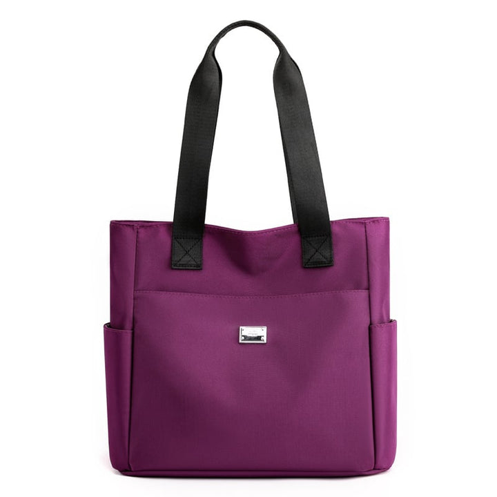 Borsa tote viola in tessuto con manici neri, tasche laterali e logo frontale. Elegante e pratica per uso quotidiano.