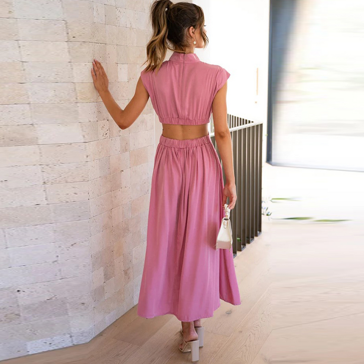 Donna in abito lungo rosa, schiena scoperta, con borsa bianca, in piedi su pavimento in legno. Moda estiva elegante, abbigliamento femminile trendy.