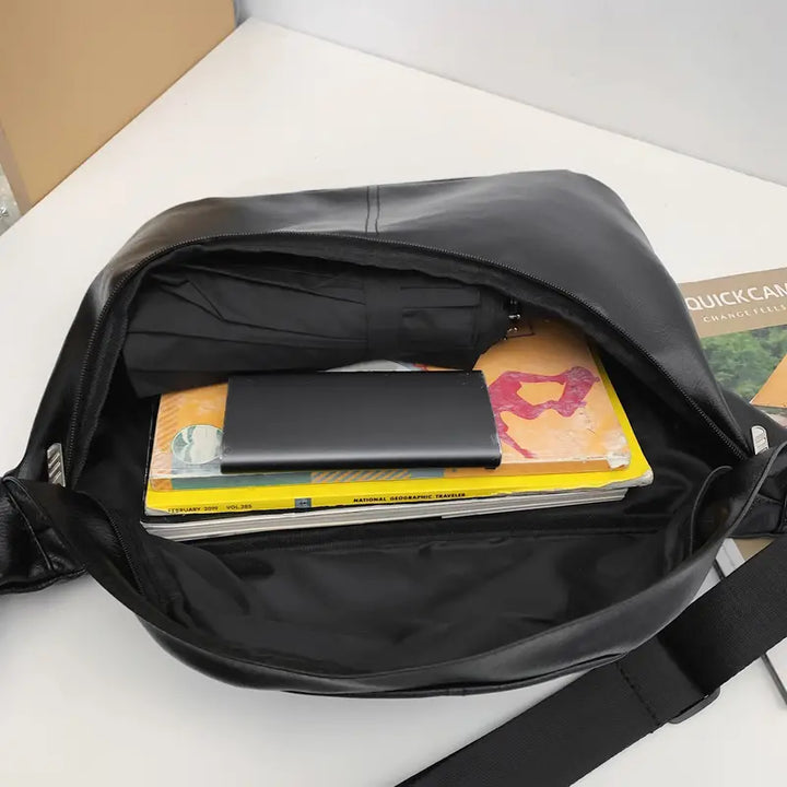 Borsa a tracolla in pelle nera aperta con ombrello, libro e power bank all'interno, su sfondo bianco. Accessori eleganti e pratici per il lavoro.