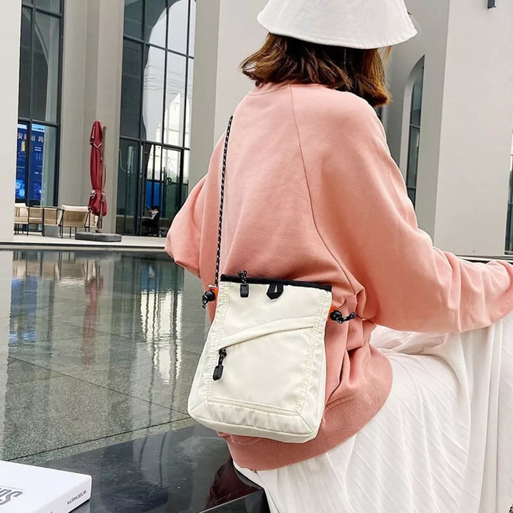 Donna con borsa a tracolla bianca, indossa felpa rosa e cappello bianco, seduta in ambiente moderno. Accessorio moda casual, stile urbano.