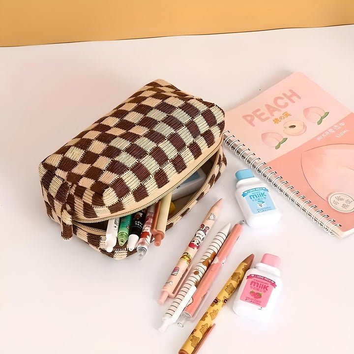 Astuccio a scacchi marrone con penne colorate, quaderno rosa "Peach" e bottigliette di latte su scrivania bianca. Accessori scolastici eleganti.