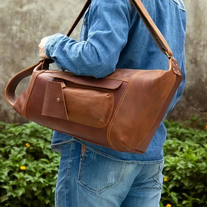 Uomo con borsa a tracolla in pelle marrone, tasca frontale con bottone, indossa giacca e jeans. Accessorio moda, stile casual, outdoor.