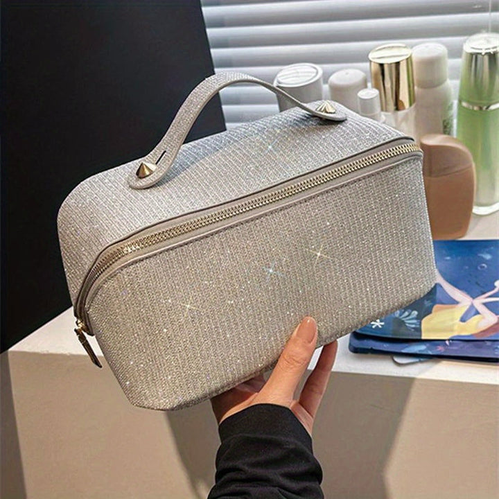 Borsa cosmetica grigia scintillante con cerniera, manico superiore e dettagli metallici, su sfondo di prodotti per la cura della pelle.