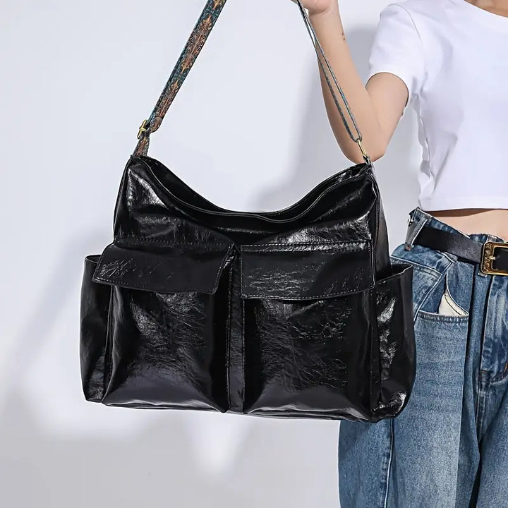 Borsa a tracolla nera in pelle lucida con tasche frontali, indossata da una donna in jeans e maglietta bianca. Accessorio moda elegante e spazioso.