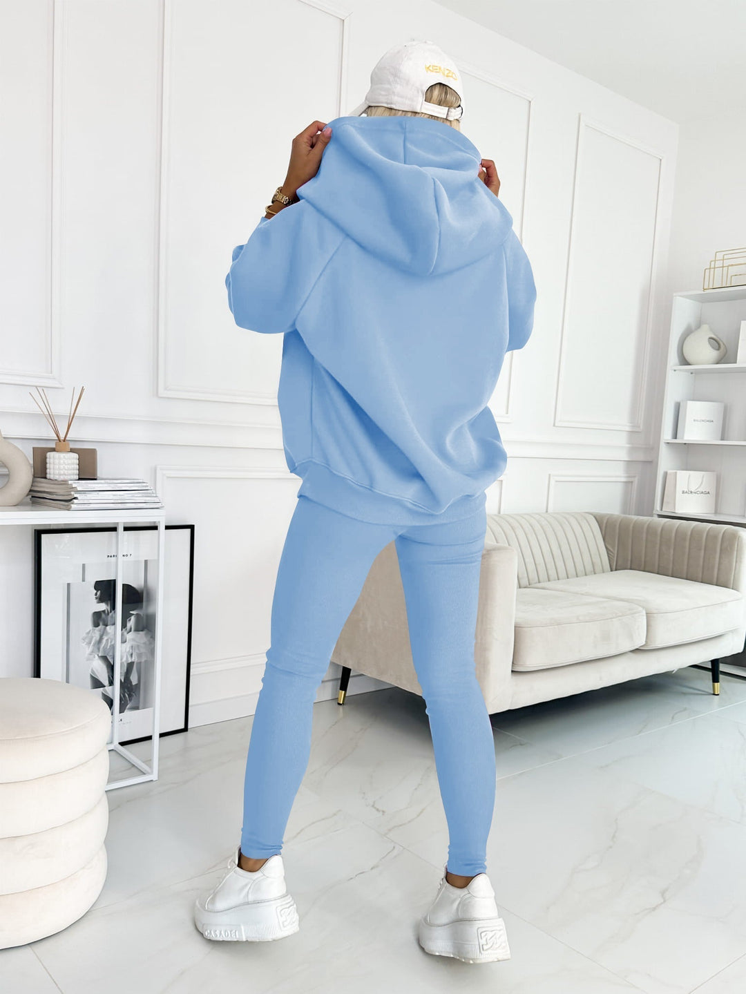 Donna in tuta sportiva azzurra, cappuccio alzato, cappellino bianco, in salotto moderno. Abbigliamento casual, moda donna, stile urbano.