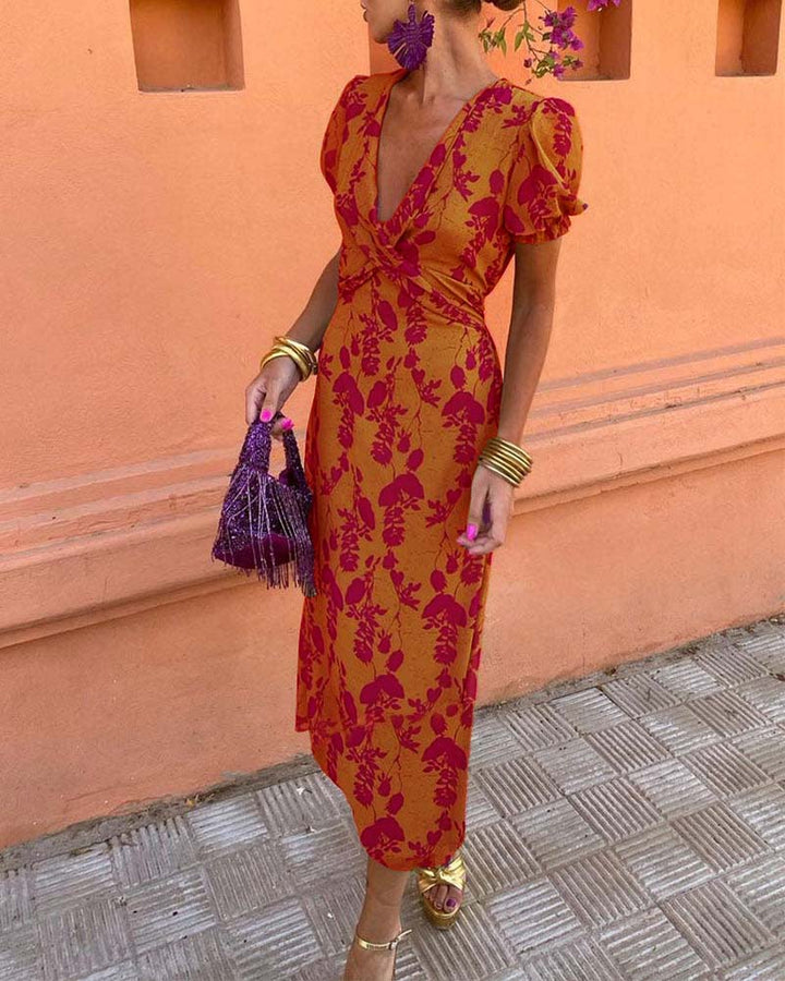 Abito lungo arancione con stampa floreale viola, maniche a sbuffo, scollatura a V. Accessori: borsa viola con frange, sandali dorati, bracciali dorati.