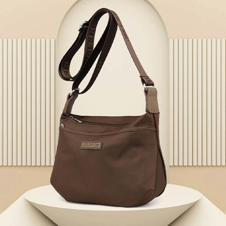 Borsa a tracolla marrone in tessuto resistente, con cerniera e logo, su sfondo beige. Accessorio moda donna, elegante e pratico.