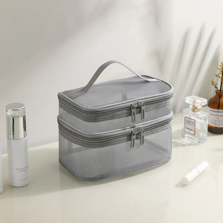 Borsa da toilette grigia a due scomparti con cerniera, manico superiore, su tavolo con profumo e cosmetici. Organizzatore da viaggio elegante.