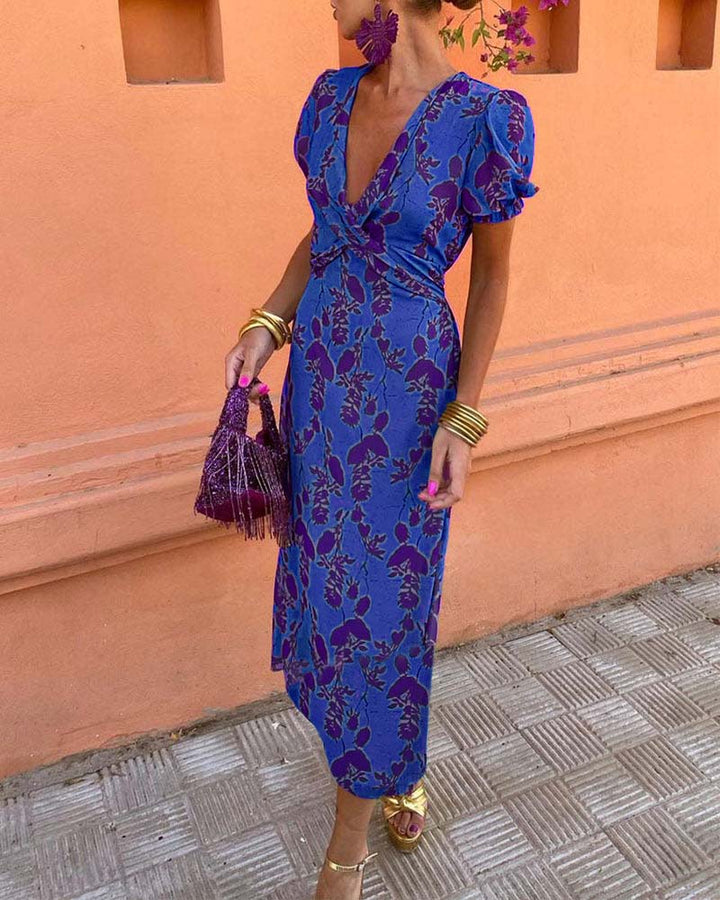 Abito lungo blu con stampa floreale viola, maniche a sbuffo, scollo a V profondo. Accessori: borsa frange viola, orecchini grandi, bracciali dorati.