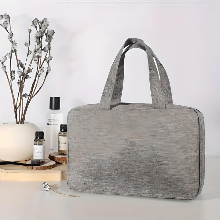 Borsa grigia elegante su sfondo bianco, accanto a flaconi di cosmetici e decorazioni floreali. Accessorio moda, design moderno, versatile.