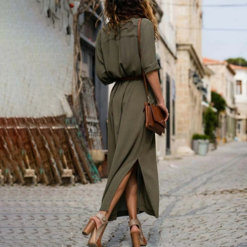 Donna con abito lungo verde oliva, sandali con tacco e borsa marrone, cammina su strada acciottolata. Moda estiva, stile casual chic.