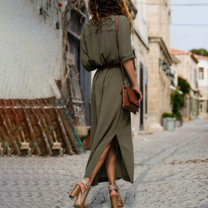 Donna con abito lungo verde oliva, sandali con tacco e borsa marrone, cammina su strada acciottolata. Moda estiva, stile casual chic.