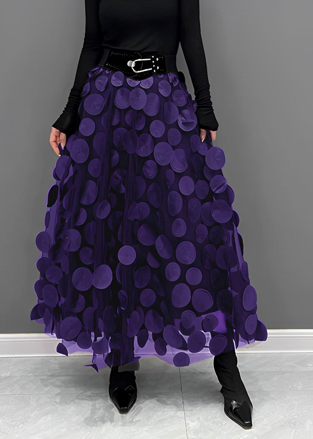 Gonna lunga viola con cerchi decorativi, cintura nera, indossata con maglia nera a maniche lunghe e stivali neri. Moda elegante donna.