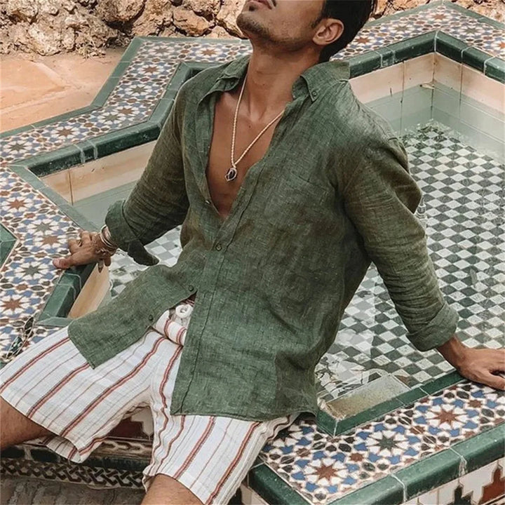 Uomo con camicia di lino verde e pantaloncini a righe bianche e marroni, seduto su fontana decorata con piastrelle geometriche. Moda estiva casual.