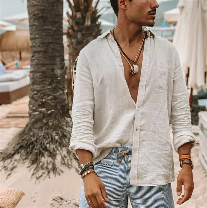 Uomo in camicia di lino bianca aperta e pantaloncini a righe blu su spiaggia tropicale, accessoriato con collane e bracciali. Moda estiva casual.