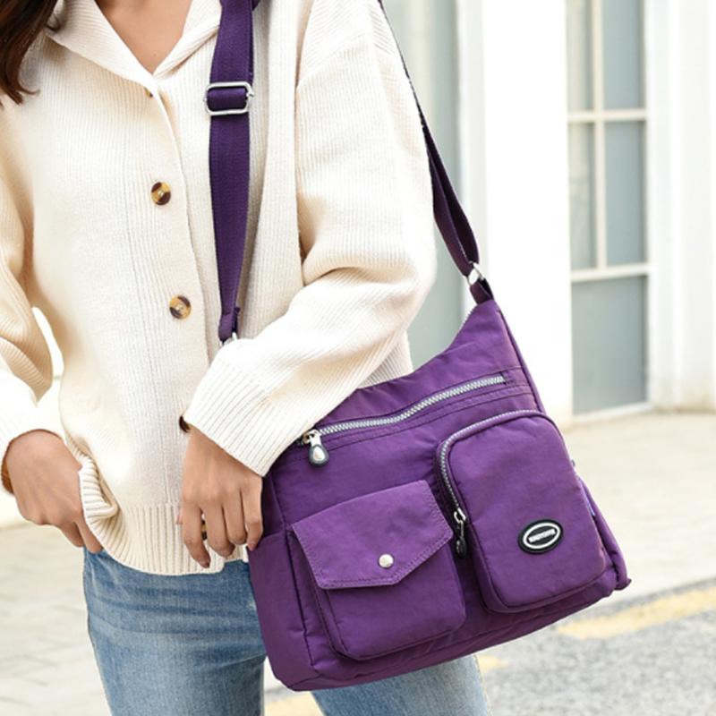 Borsa a tracolla viola in nylon con tasche multiple, indossata da una persona in cardigan bianco e jeans. Accessorio moda casual e pratico.