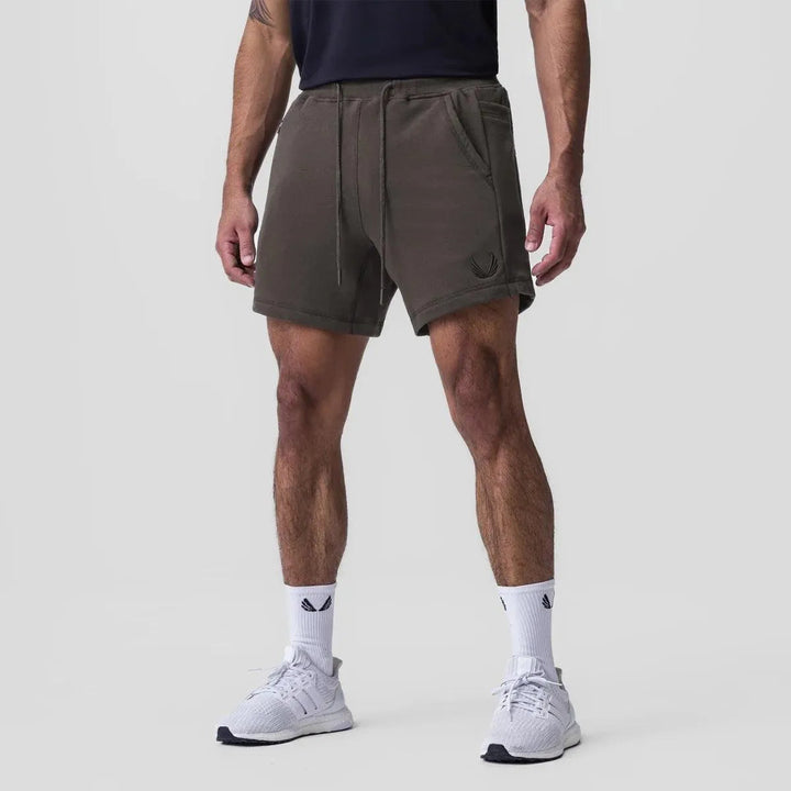 Shorts sportivi uomo in cotone verde oliva, con tasche laterali e coulisse, indossati con scarpe da ginnastica bianche. Ideali per allenamento.