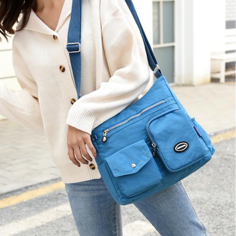 Borsa a tracolla blu in tessuto, con tasche multiple e cerniere, indossata da una persona in jeans e maglione bianco. Accessorio moda casual.