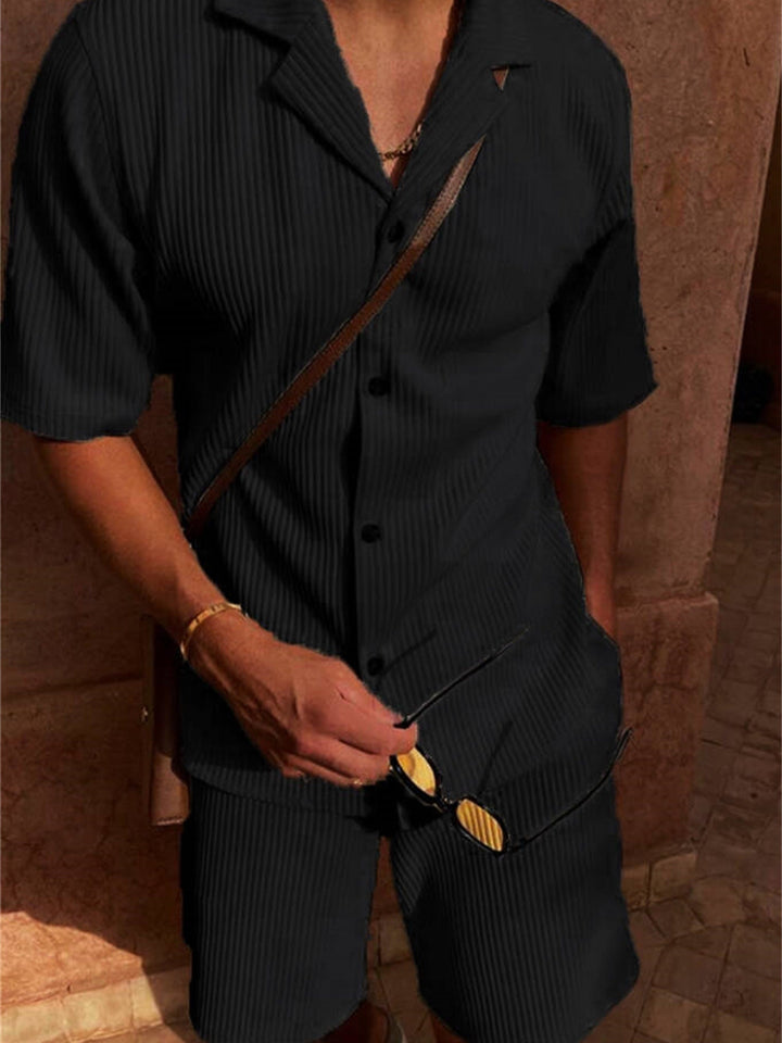 Uomo in completo estivo nero a righe, camicia a maniche corte e pantaloncini coordinati, con occhiali da sole e borsa a tracolla marrone.