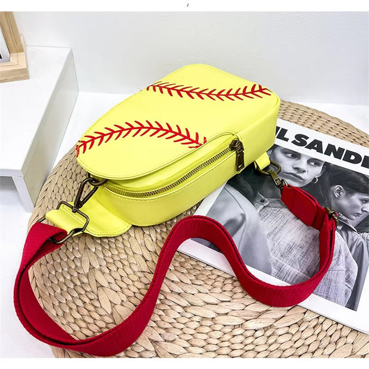Borsa a tracolla gialla a forma di palla da baseball con cuciture rosse, cinturino rosso regolabile, su tavolo bianco. Accessorio moda sportivo.