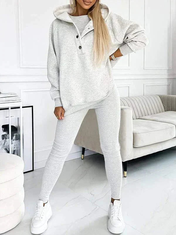 Donna in tuta sportiva grigia, felpa con cappuccio e pantaloni abbinati, in salotto moderno. Abbigliamento casual, moda donna, comfort.
