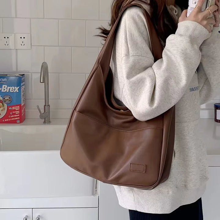 Borsa a spalla in pelle marrone su sfondo cucina, indossata da persona con felpa grigia. Accessorio moda elegante e pratico per uso quotidiano.