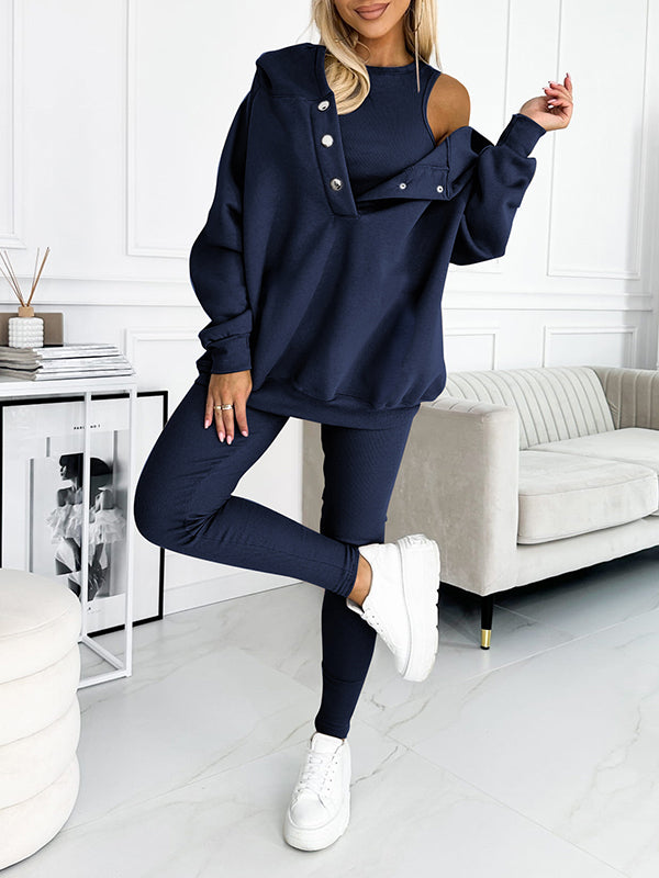 Donna in tuta sportiva blu scuro, design asimmetrico, spalla scoperta, abbinata a sneakers bianche. Outfit casual elegante per moda autunnale.