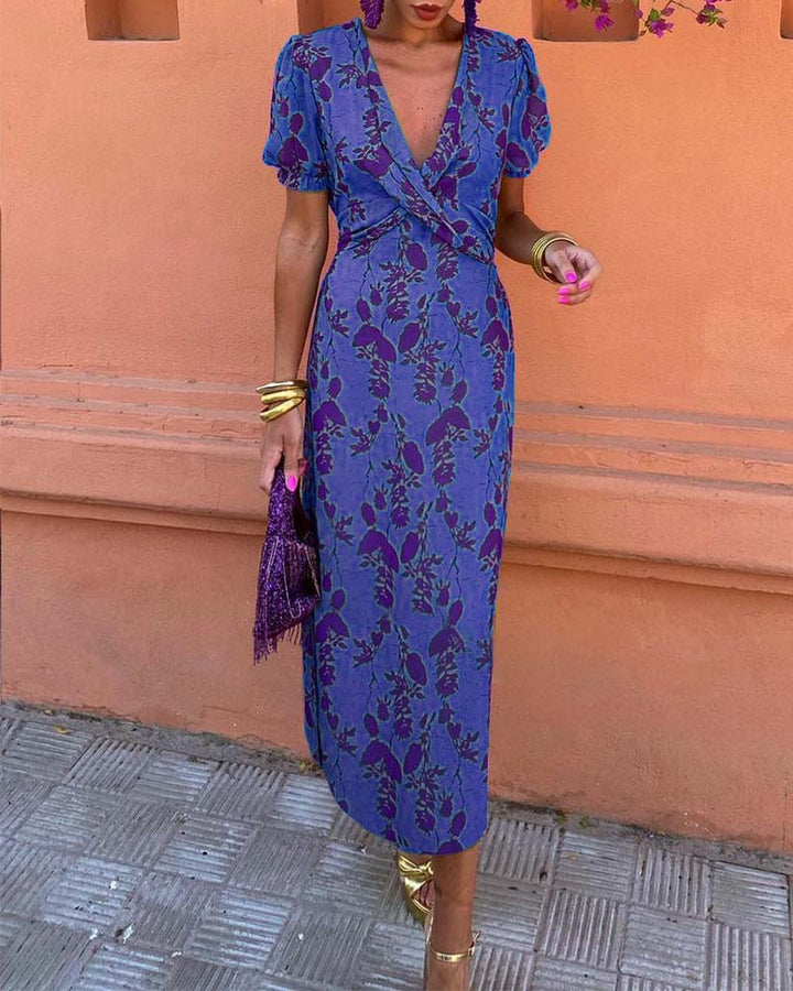 Abito lungo floreale blu e viola con scollo a V, maniche corte e accessori dorati. Perfetto per moda estiva elegante e occasioni speciali.