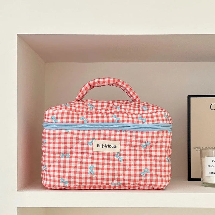 Borsa da toilette a quadretti rossi e bianchi con cerniera azzurra, manico superiore e motivo floreale, su mensola bianca.