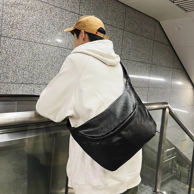 Uomo con cappellino beige e felpa bianca indossa una borsa a tracolla nera in pelle, appoggiato a una ringhiera in metropolitana.