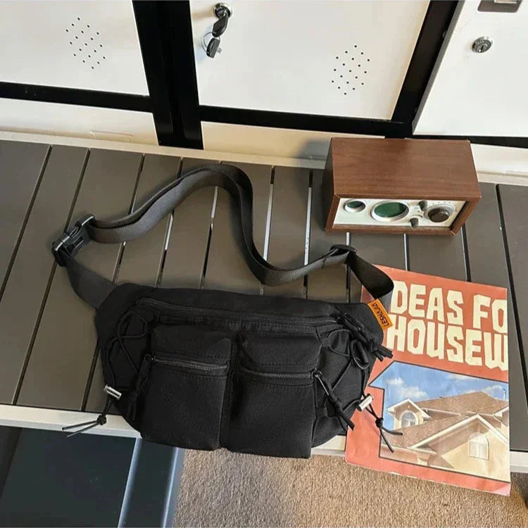 Marsupio nero con tasche multiple su tavolo grigio, accanto a rivista "Ideas for Housew" e radio vintage. Accessorio moda pratico e versatile.