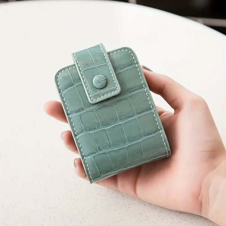 Portafoglio donna in pelle verde con texture coccodrillo, chiusura a bottone, design compatto, elegante e moderno, tenuto in mano su sfondo chiaro.