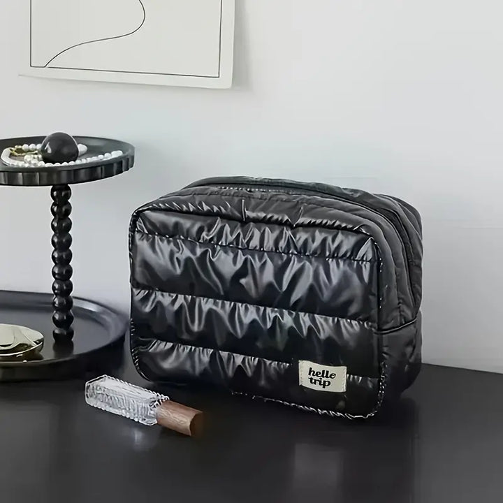 Borsa da toilette nera trapuntata su tavolo moderno, con etichetta "hello trip", accanto a vassoio con perle e profumo. Design elegante e funzionale.