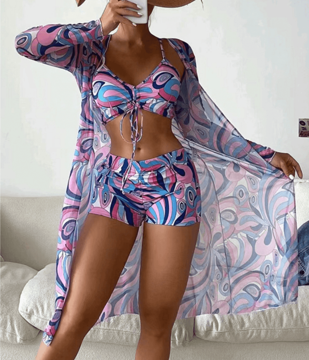 Donna in costume da bagno fantasia psichedelica rosa e blu con copricostume coordinato, posa su divano bianco. Moda estiva, abbigliamento mare.