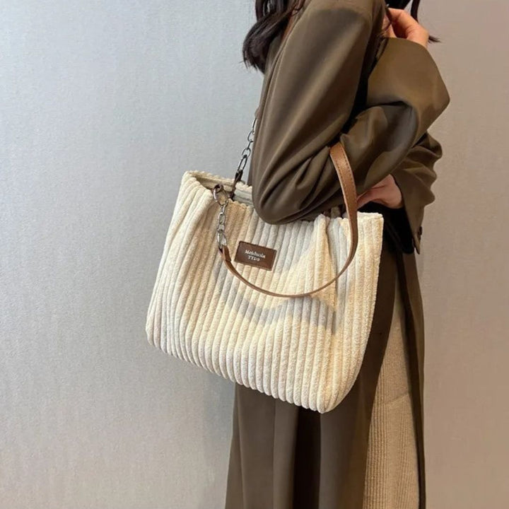 Donna con cappotto marrone indossa borsa a spalla in tessuto beige a coste, manico in pelle marrone, elegante e moderna. Accessorio moda autunno.