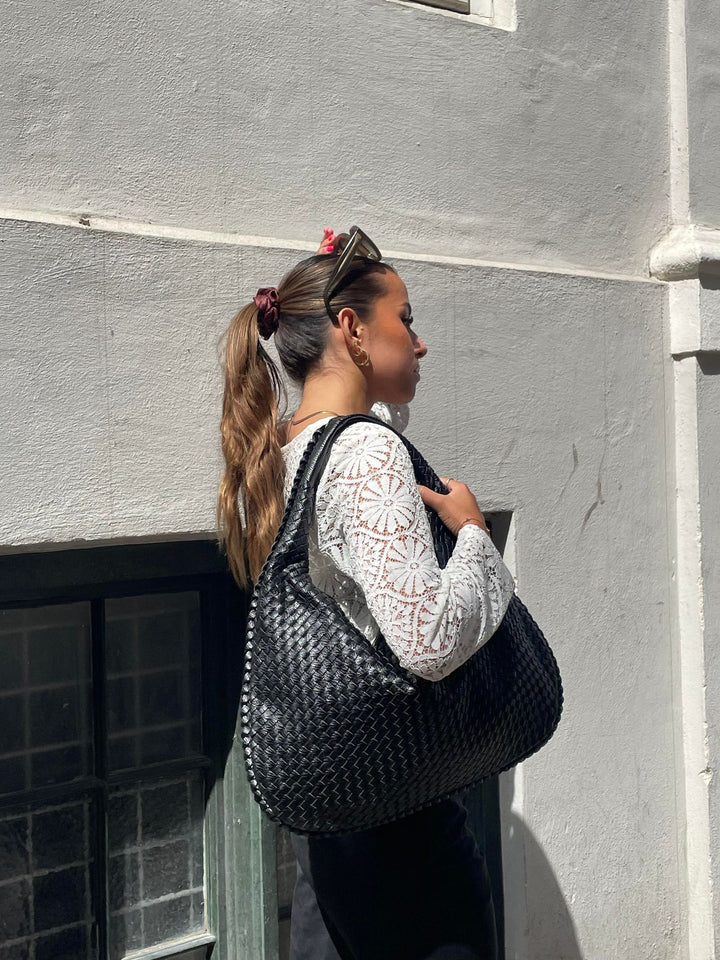 Donna con borsa intrecciata nera, indossa maglia bianca a maniche lunghe con motivo floreale. Moda urbana, accessori eleganti, stile casual chic.