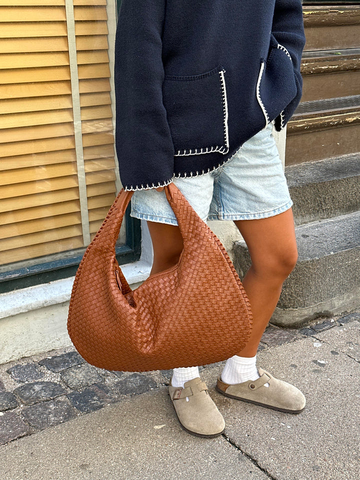 Donna con borsa a spalla intrecciata marrone, indossa giacca blu e pantaloncini di jeans. Scarpe beige, look casual urbano.