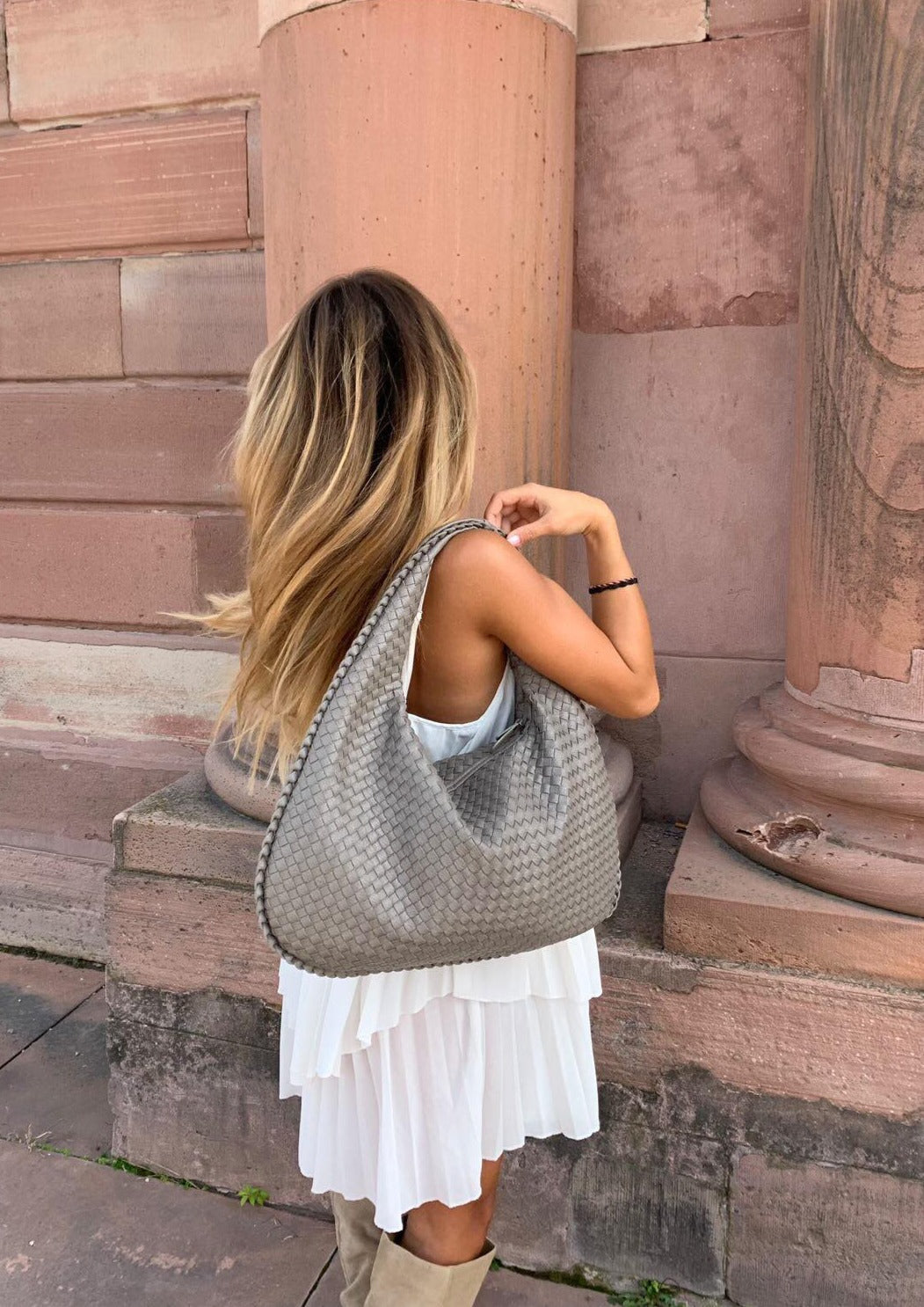 Donna con borsa a spalla intrecciata grigia, abito bianco e stivali beige, davanti a colonne di pietra. Moda autunnale elegante e chic.