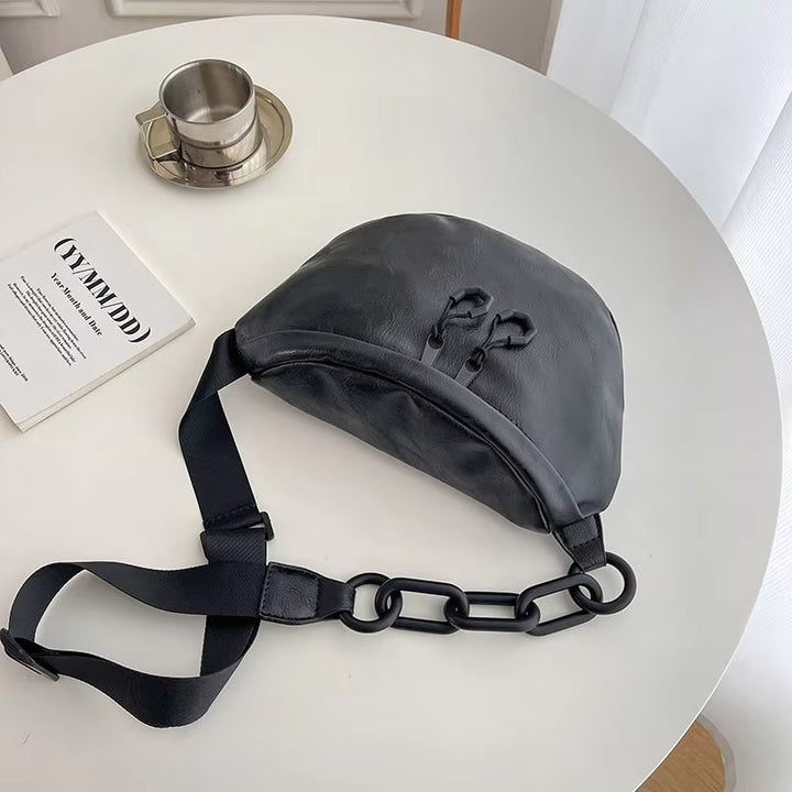 Marsupio nero in pelle su tavolo bianco, con catena decorativa e cinghia regolabile. Accessorio moda unisex, stile urbano e contemporaneo.