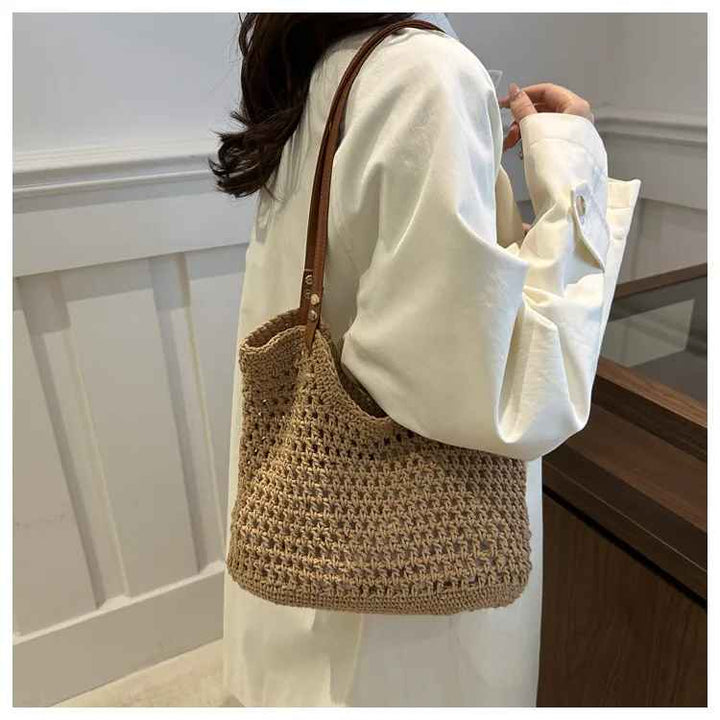 Donna con borsa a tracolla in paglia intrecciata, stile casual, manico in pelle marrone, indossa camicia bianca. Accessorio moda estivo elegante.