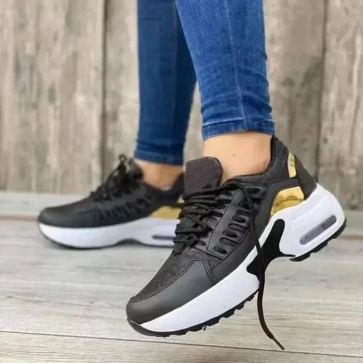 Scarpe da ginnastica nere e oro con suola bianca, indossate da una persona su pavimento in legno. Sneakers moda, casual, comode.