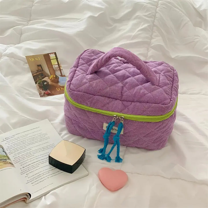 Borsa cosmetica viola trapuntata su letto bianco, con cerniera verde e nappine blu, accanto a libro aperto e cuore rosa. Accessori da viaggio eleganti.
