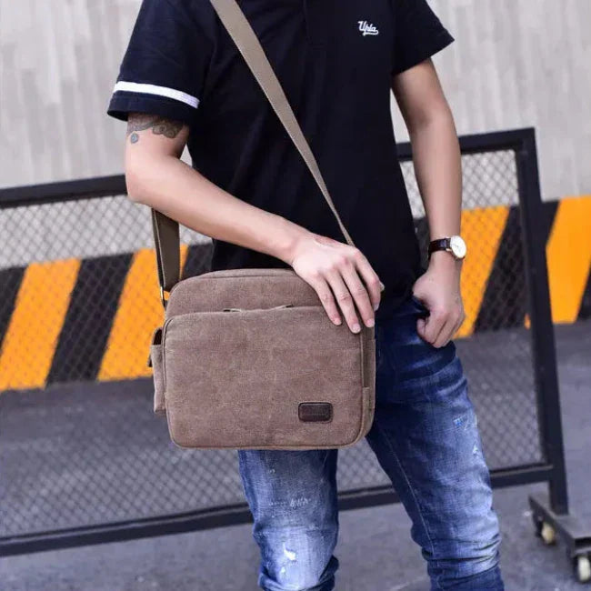 Uomo con borsa a tracolla marrone in tela, indossa jeans e maglietta nera. Accessorio moda casual, ideale per lavoro o viaggio.