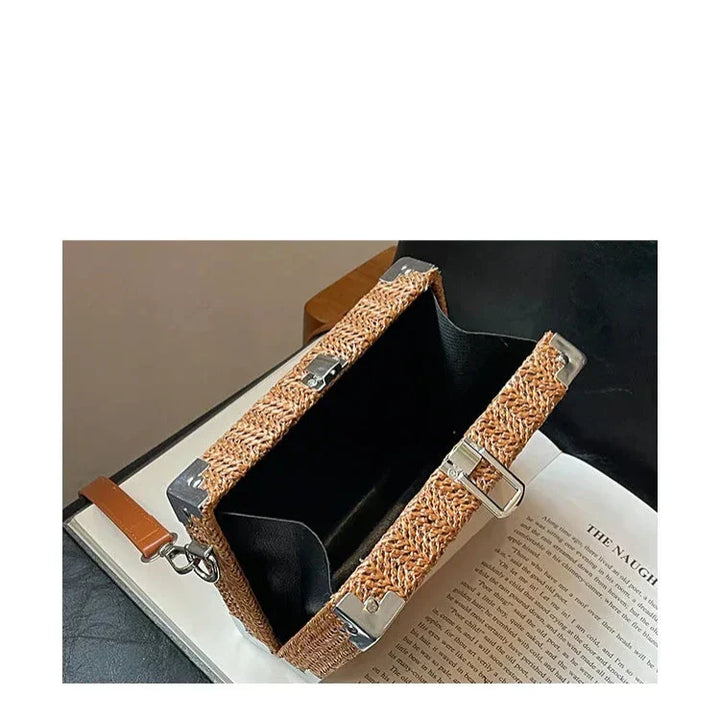 Borsa a tracolla in paglia intrecciata con dettagli in metallo, design rettangolare, aperta su libro, stile estivo elegante e moderno.