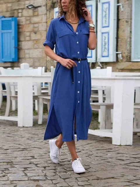 Donna con abito camicia blu, cintura nera, scarpe da ginnastica bianche, cammina su strada acciottolata. Moda casual estiva, look urbano elegante.