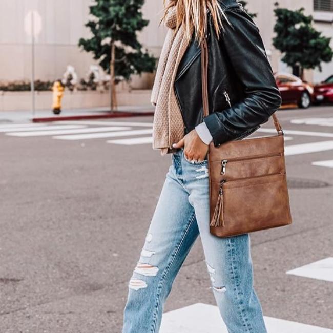 Donna con giacca in pelle nera, jeans strappati e borsa a tracolla marrone in pelle, attraversa la strada. Stile casual urbano, moda autunnale.