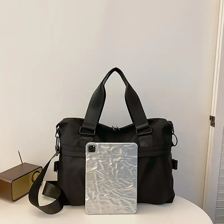 Borsa a tracolla nera elegante con manici, accanto a un tablet argento. Design moderno, ideale per lavoro e viaggio. Accessori moda unisex.