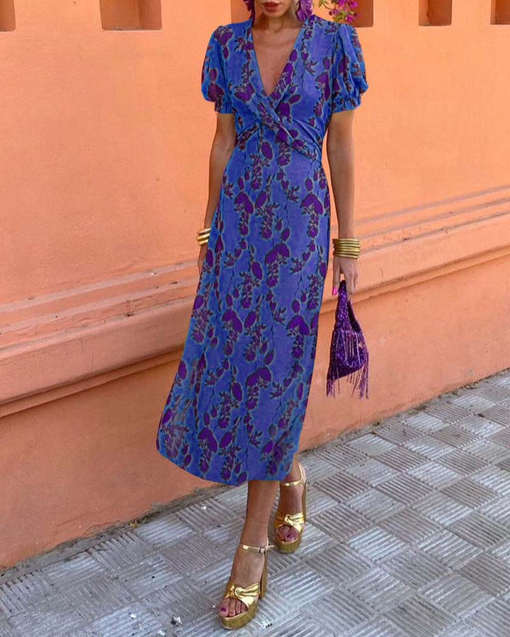 Abito lungo floreale blu e viola con maniche a sbuffo, borsa frangiata viola, sandali dorati. Donna su sfondo arancione. Moda estiva elegante.