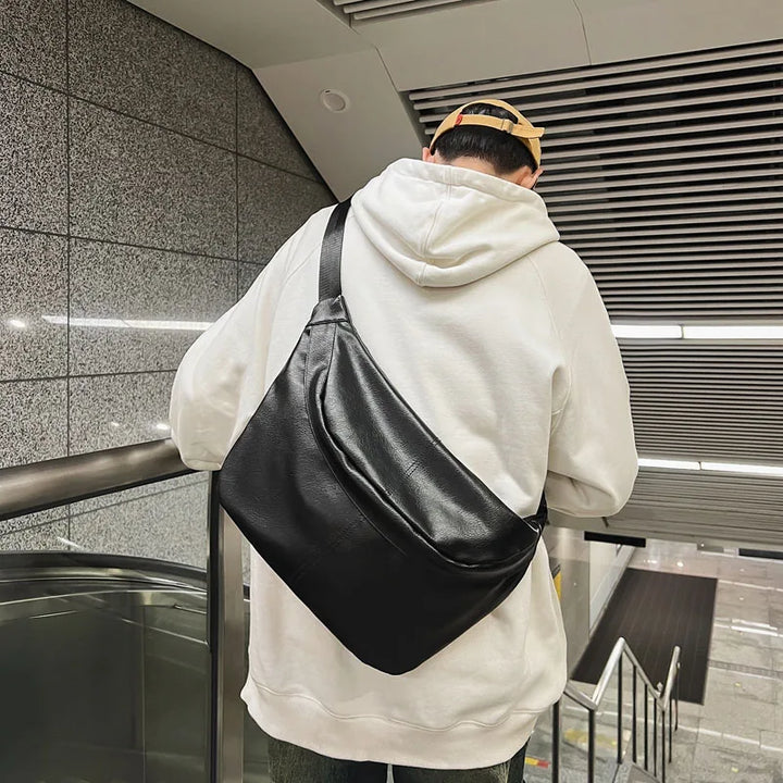 Uomo con felpa bianca e cappellino beige indossa borsa a tracolla nera in pelle, stile urbano, su scala mobile in metropolitana moderna.