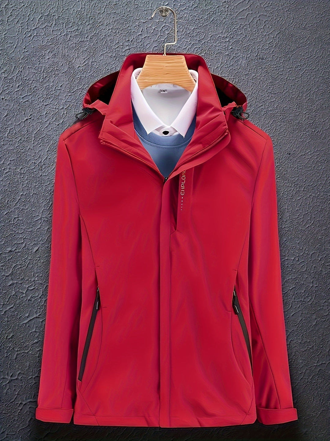 Giacca rossa impermeabile con cappuccio, appesa su gruccia, sopra camicia bianca e maglione blu. Abbigliamento outdoor elegante e funzionale.